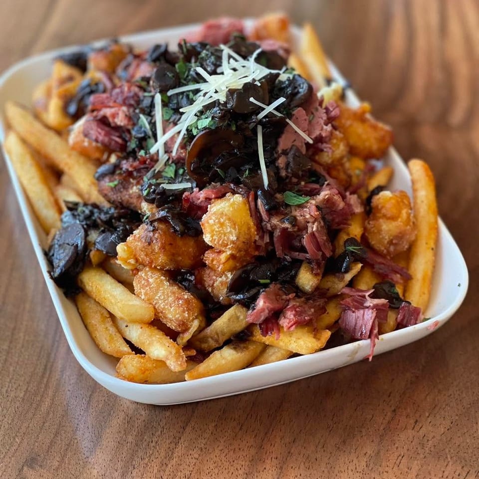 A white bowl of poutine with fries, cheese curds, shredded meat, mushrooms, and cheese.