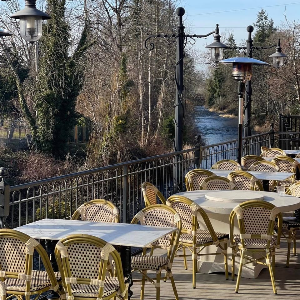 Outdoor dining patio with wicker chairs and marble tables overlooking a river and trees.