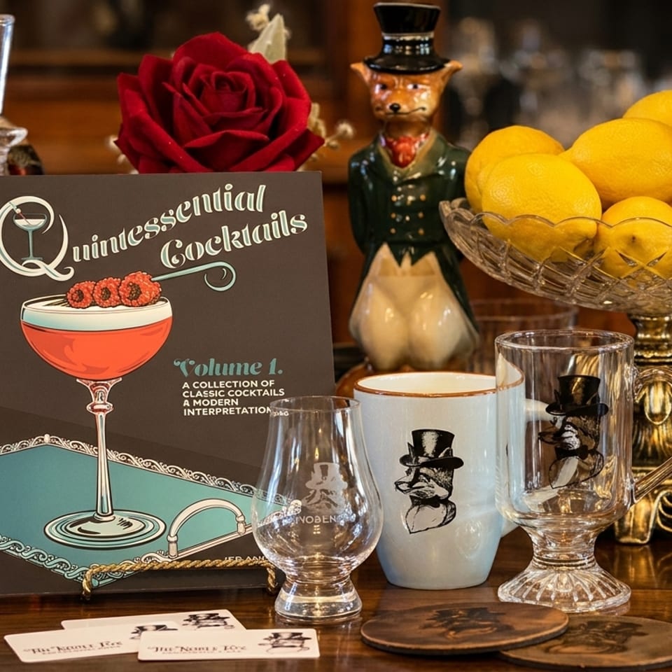 A cocktail recipe book, decanter, lemons, and branded glassware on a wooden table.
