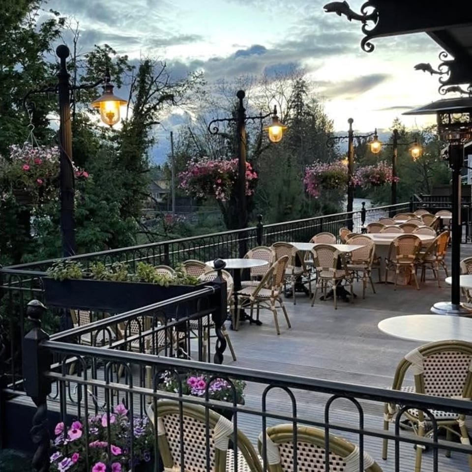 An empty outdoor restaurant patio with tables and chairs, overlooking a river at dusk.