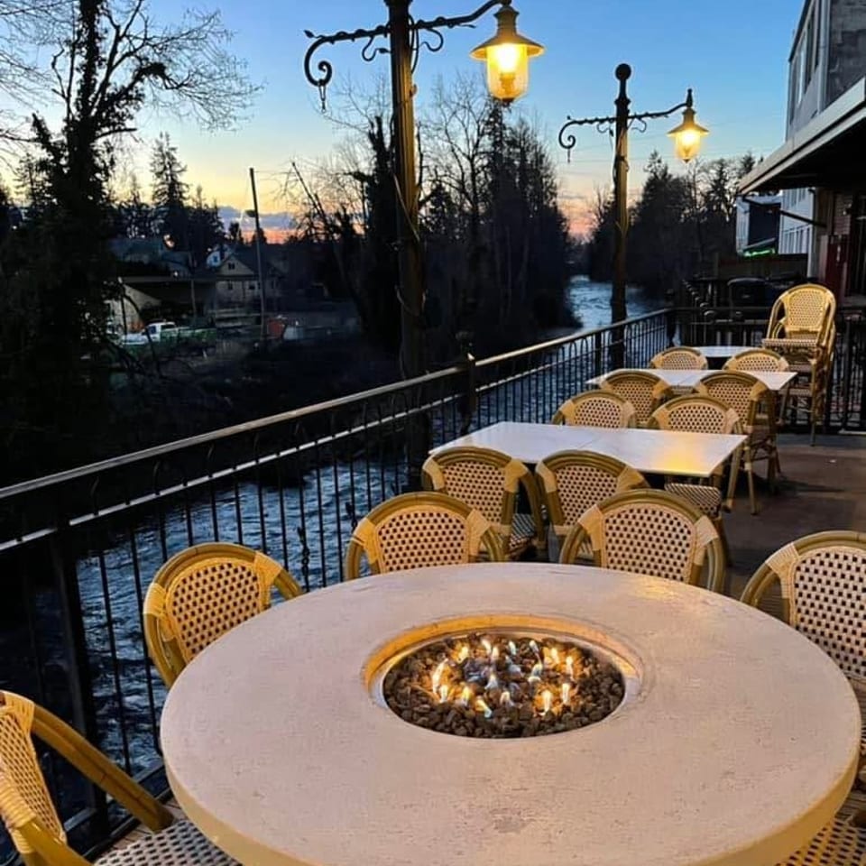 Outdoor restaurant patio with a fire pit table and river view at dusk.