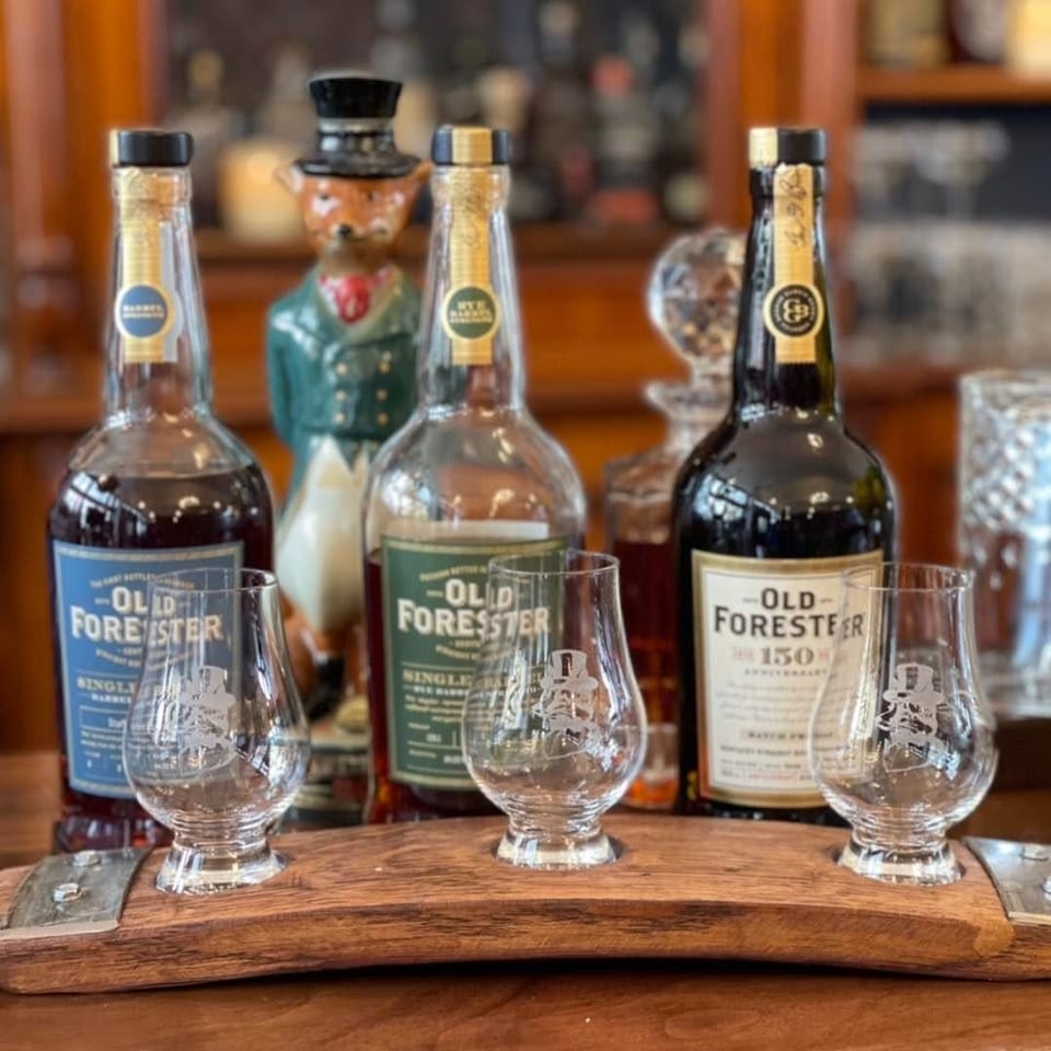 Three bottles of Old Forester whiskey with three branded tasting glasses on a wooden barrel stave.